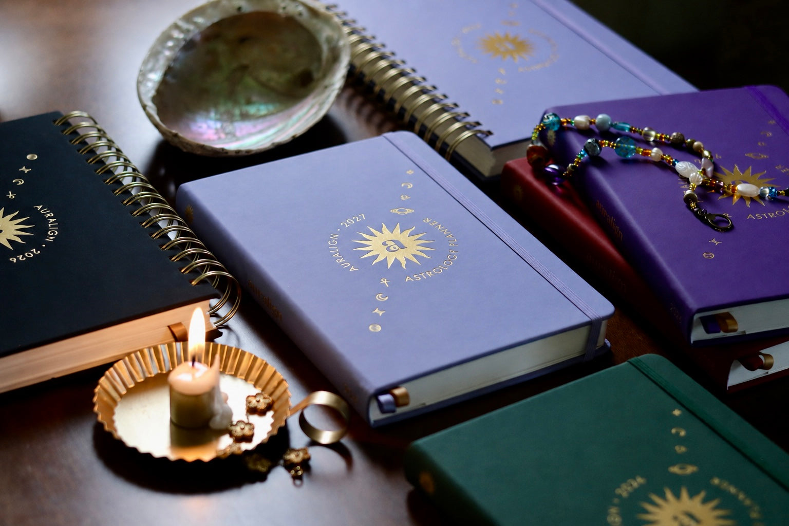 A styled arrangement of several hardcover notebooks or journals with gold sunburst logos, including a lavender one in the center, a purple one on the right, a dark green one at the bottom, and a black one on the left. A small lit candle in a gold dish, an iridescent shell, and a beaded bracelet rest on the books against a dark wooden surface.