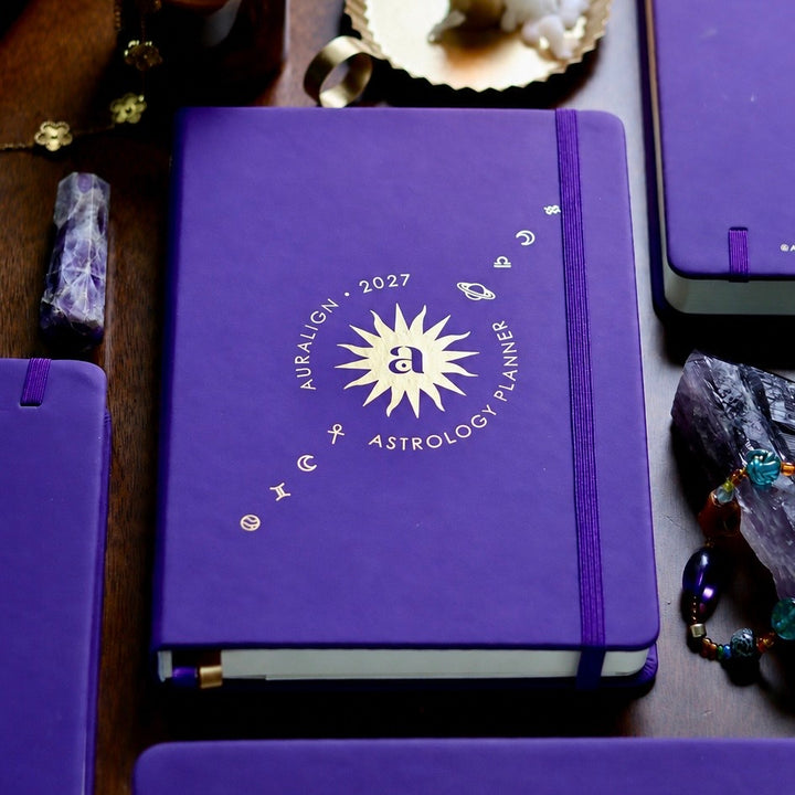 A purple hardcover astrology planner with a gold sunburst and zodiac symbols on the front, displayed on a dark wooden surface with crystals and other matching planners around it.