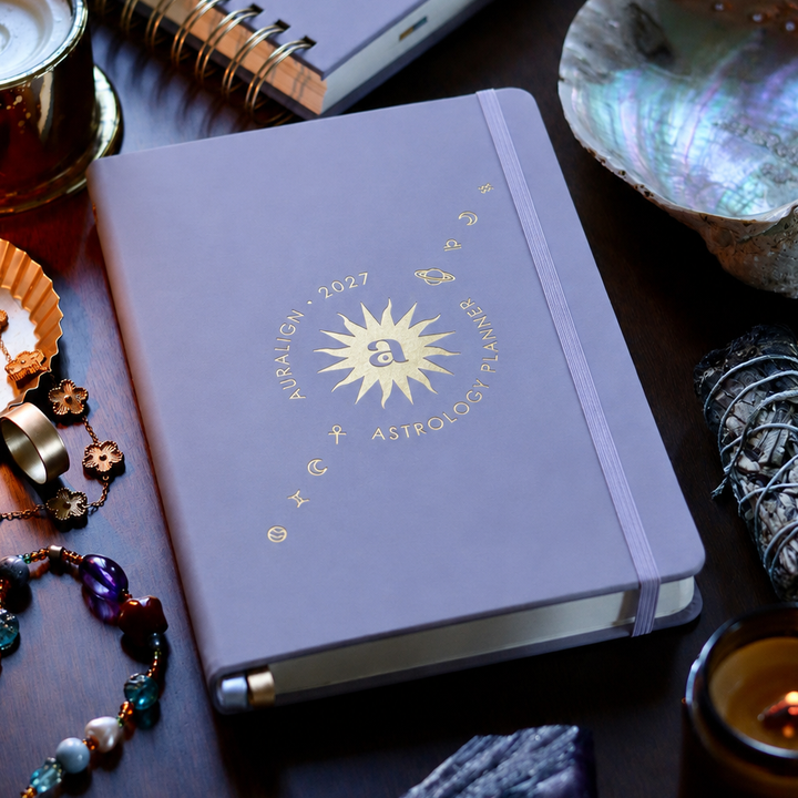 A lavender-purple hardcover journal with gold foil astrological designs, styled on a dark wooden table with crystals, a candle, and other spiritual accessories.
