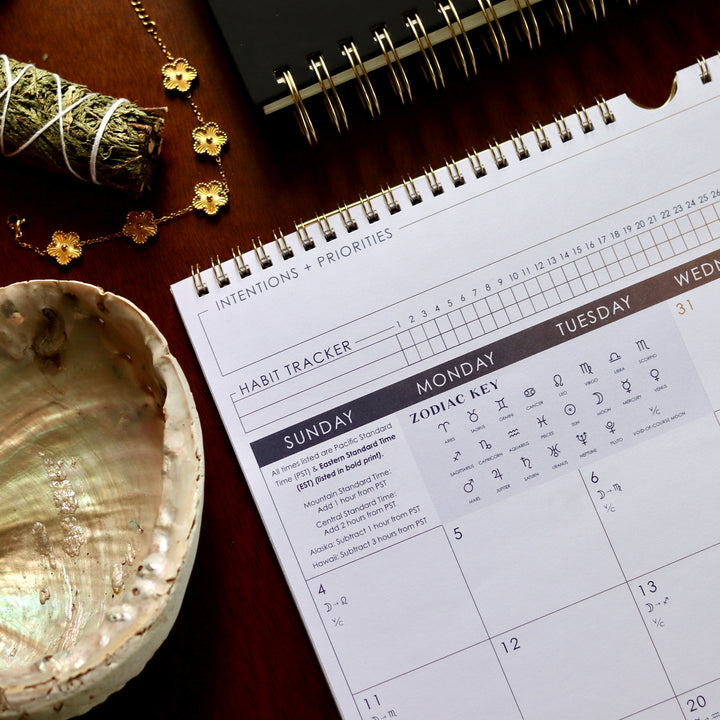 Calendar with habit tracker and zodiac key on a wooden surface with a shell and flowers.