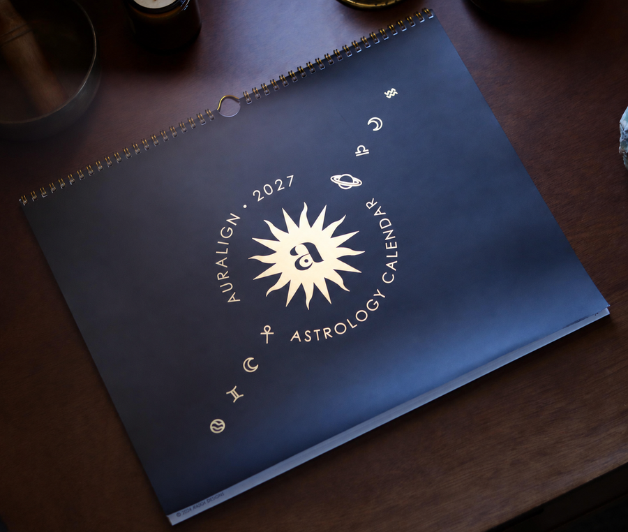 A spiral-bound 2027 astrology calendar titled 'Auralign Astrology Calendar' with a gold sunburst and zodiac symbols on a dark navy cover, photographed on a dark wooden tabletop.