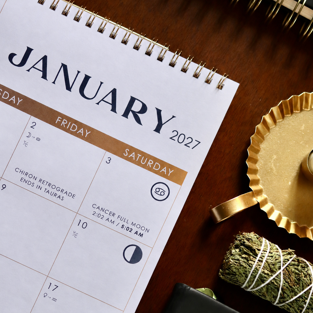 A spiral-bound January 2027 calendar page laid on a dark wooden surface, surrounded by styled props including a gold tray, a bundle of dried herbs, and part of a notebook.