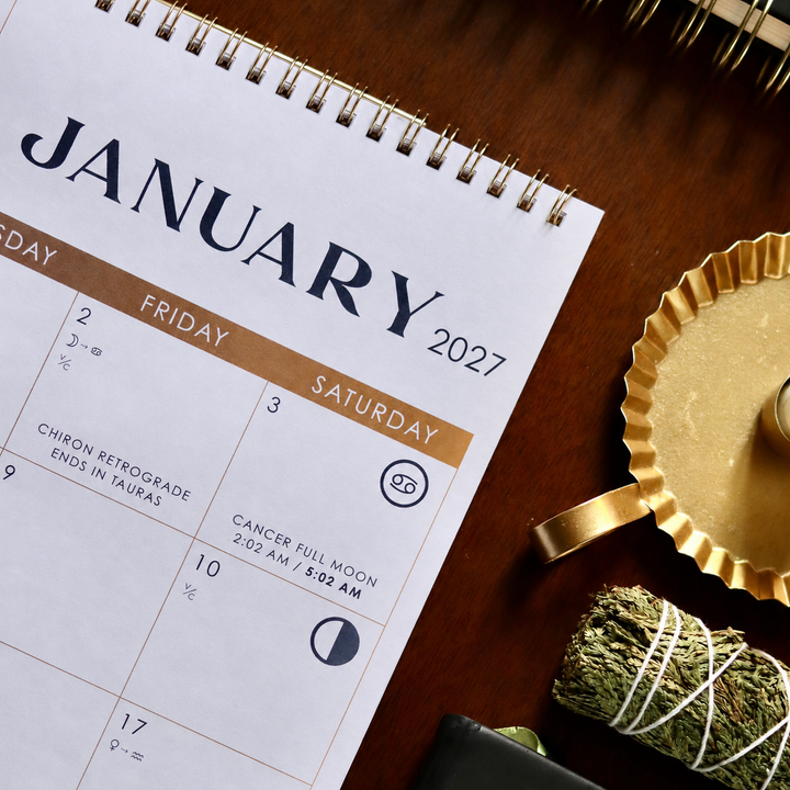 A spiral-bound January 2027 calendar page laid on a dark wooden surface, surrounded by styled props including a gold tray, a bundle of dried herbs, and part of a notebook.
