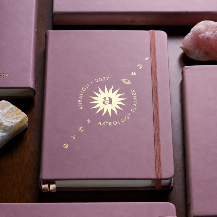 A dusty mauve purple hardcover journal titled 'AIPALION 2027 ASTROLOGY PLANNER' with gold foil lettering and a gold sunburst emblem on the front cover. The journal is closed with a matching elastic band and is displayed on a dark wooden surface alongside other matching journals and crystal stones.
