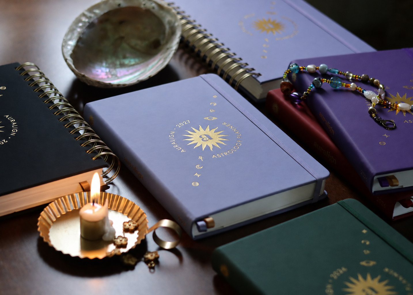 A styled flat-lay of several astrology-themed notebooks and journals with gold sunburst designs, arranged on a dark wooden surface alongside a small burning candle, a shell dish, and a beaded bracelet.
