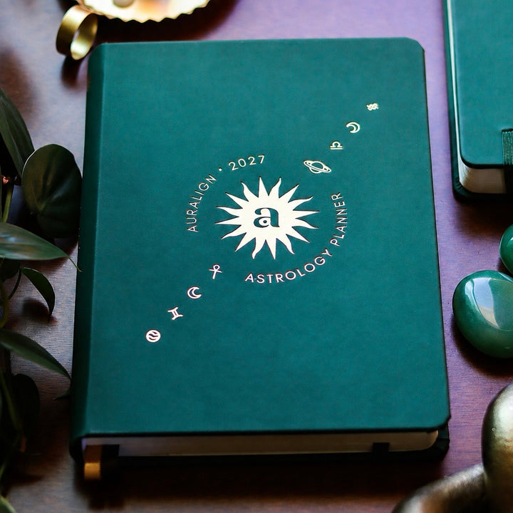 A dark teal astrology planner with a white sunburst and zodiac symbols on the cover, styled on a dark wooden surface with crystals and other notebooks.